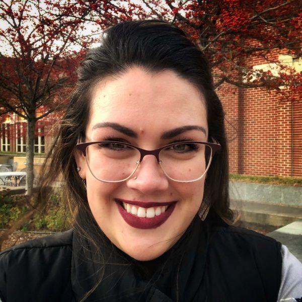 Photo of a woman smiling while outdoors. She is dressed warmly for fall weather and wears glasses.